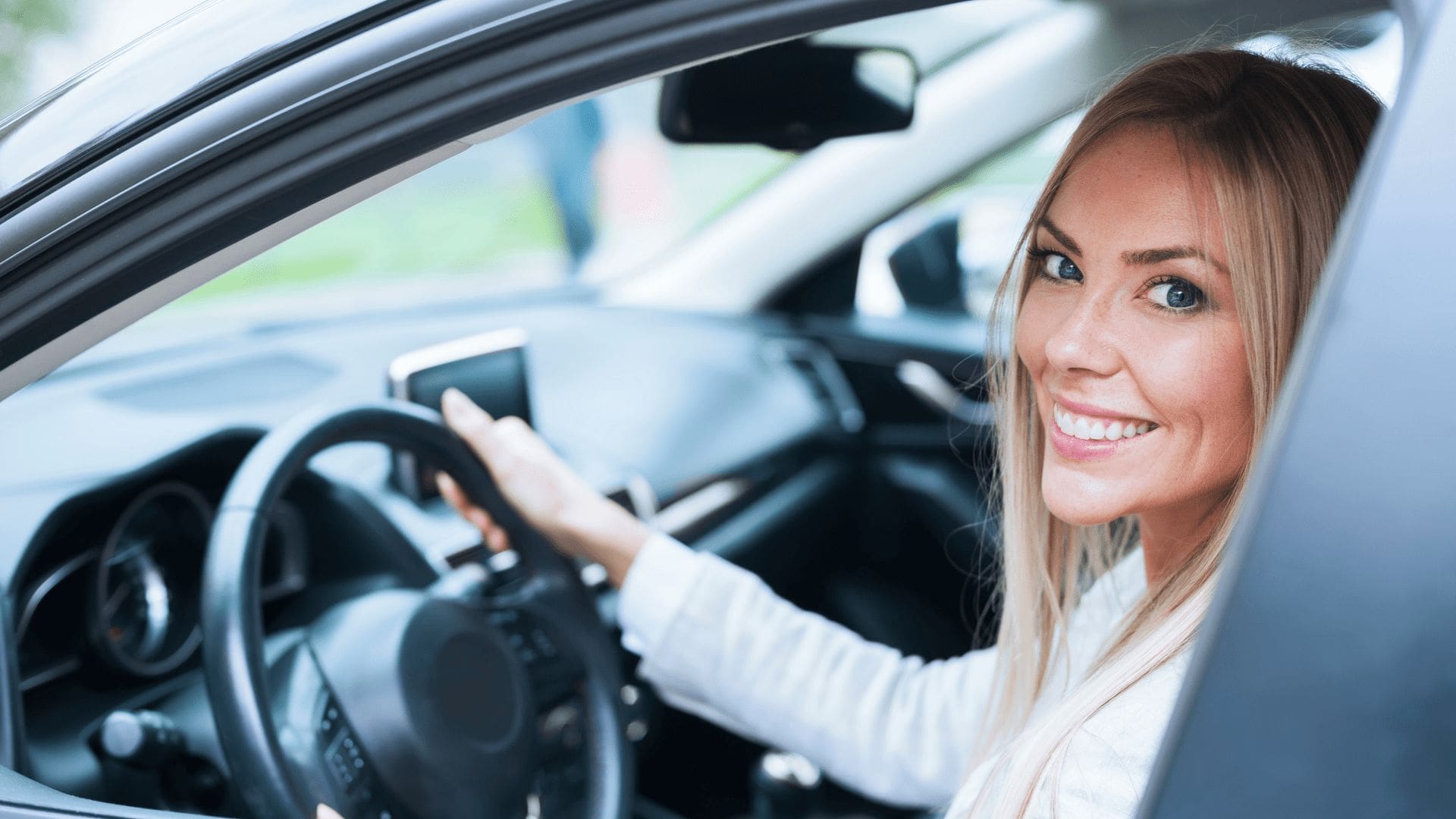 En blond vit kvinna sitter i en bil och ler.