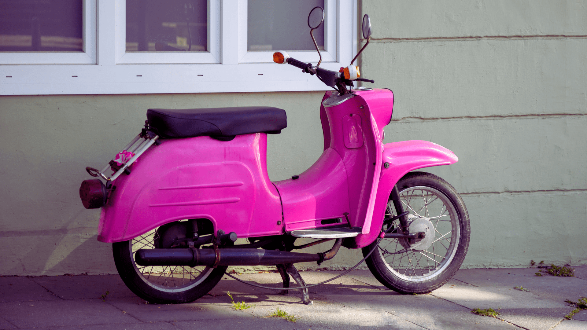 En rosa moped parkerad vid en grå vägg med vita fönster.
